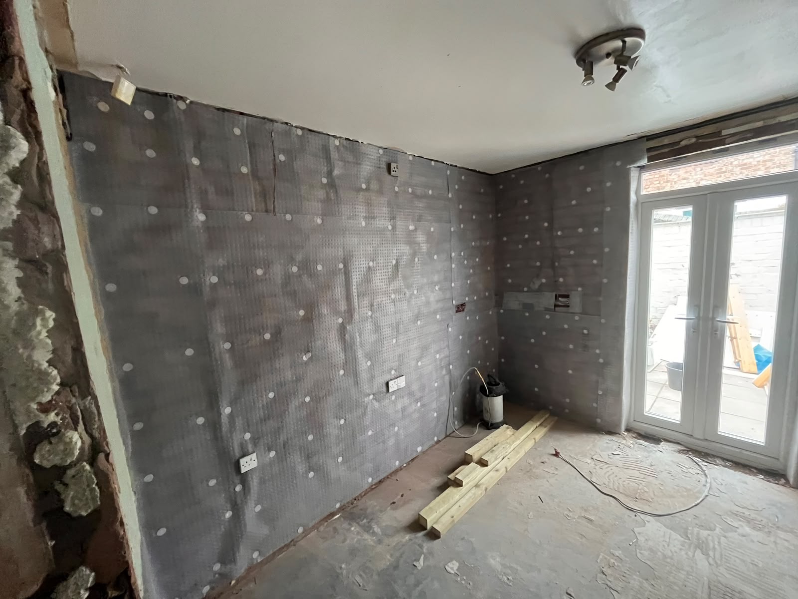 Grey damp-proof membrane with white fixing plugs covering interior masonry walls, patio doors beyond
